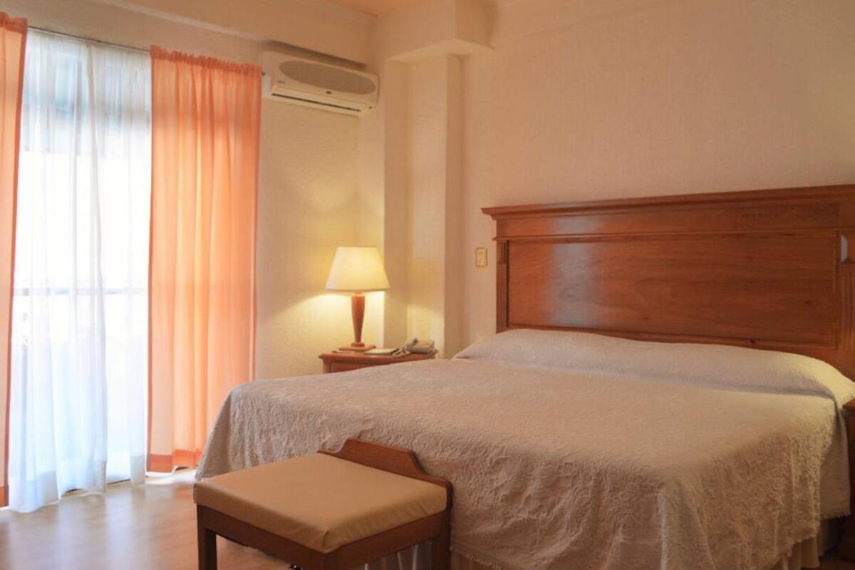 A bedroom with a wooden bed, a bedside table with a lamp, a window with curtains, and a bench.