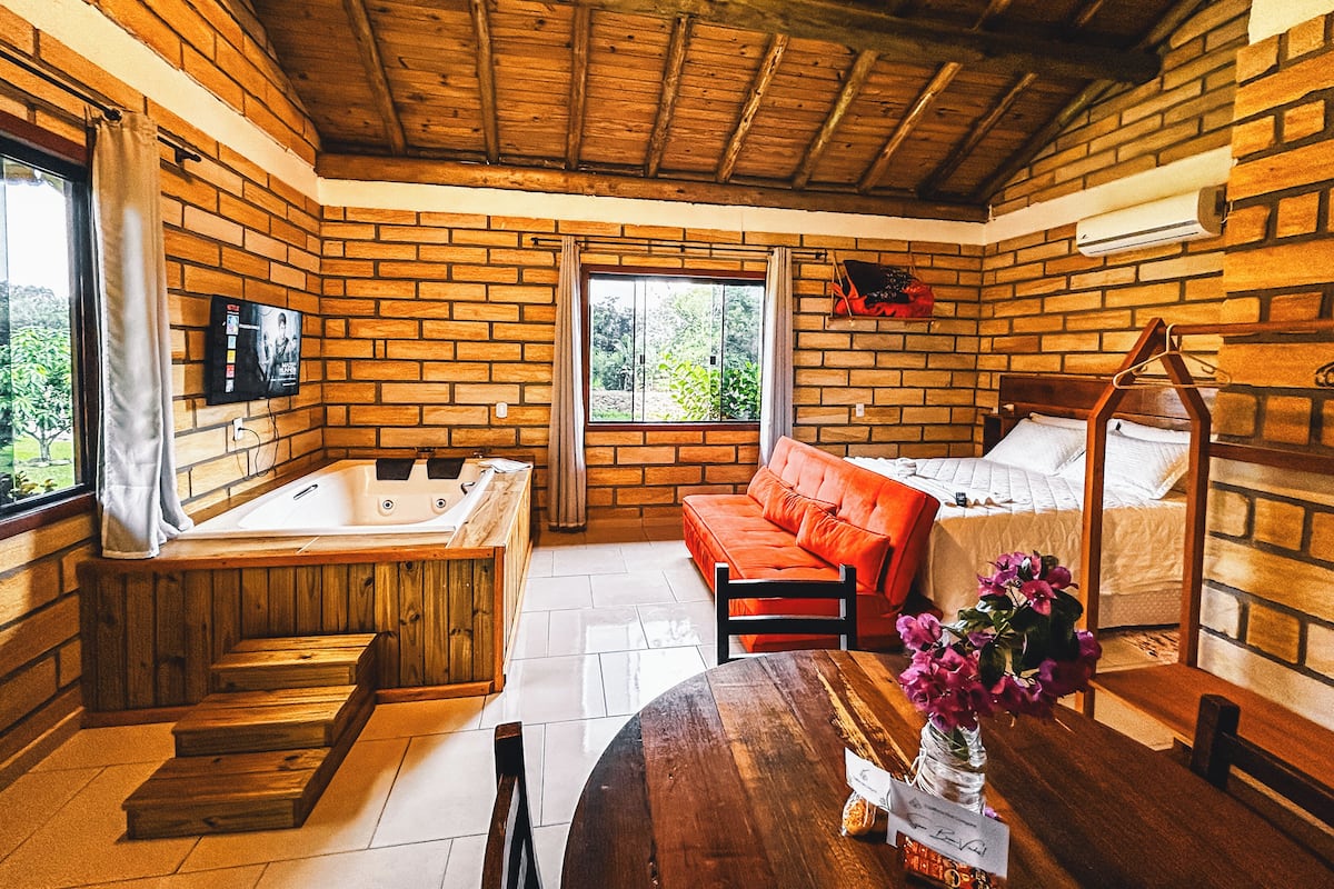 Um quarto aconchegante com teto de madeira, uma janela grande, uma banheira de madeira, um sofá vermelho, uma mesa de madeira com um vaso de flores e uma cama com cabeceira de madeira.