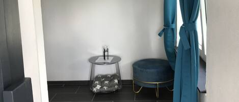 Un baño con piso de baldosas oscuras, una silla azul redonda, una pequeña mesa redonda con una silla, y tres obras de arte colgadas en la pared.