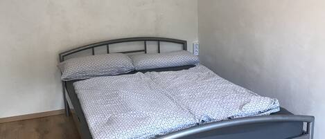 Una cama individual con estructura metálica y ropa de cama estampada en una habitación con suelo de madera y paredes lisas.