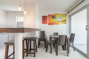 Un comedor con una mesa y sillas de madera, un ventanal con persianas, y tres cuadros con paisajes al atardecer en la pared.