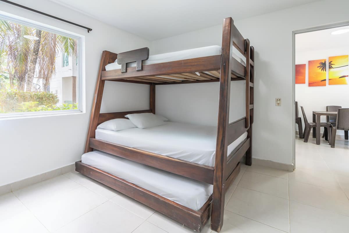 A bunk bed with a wooden frame, white bedding, and a window with a view of palm trees.