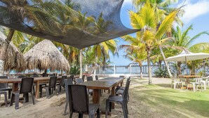 An outdoor dining area with tables and chairs, a thatched roof, and palm trees. - Cabañas Aqua Blue Coveñas (Coveñas)