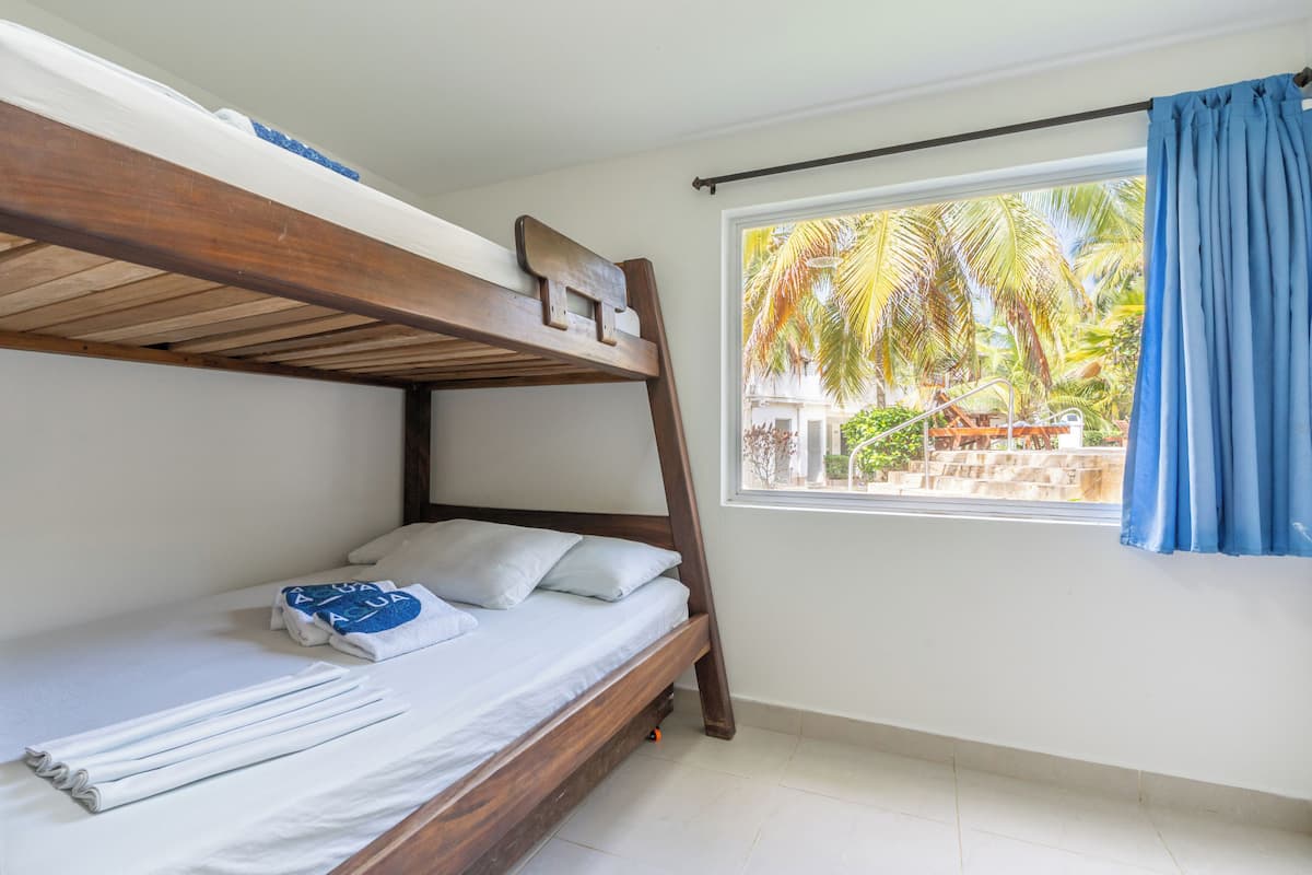 A bunk bed with a wooden headboard, a window with a view of palm trees, and a blue curtain.