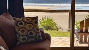 Un canapé marron agrémenté d’un coussin à motifs, une fenêtre donnant sur une plage et une chaise en bois à l’extérieur.