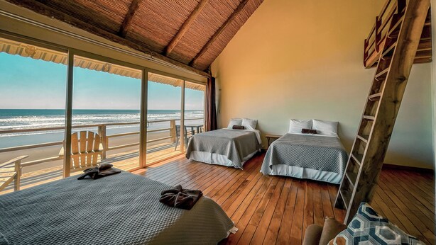 Une chambre avec deux lits, un plancher en bois, une vue sur l’océan et un toit de chaume.