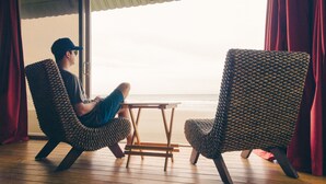 Une pièce avec un plancher en bois, deux chaises à motifs, une petite table en bois et une vue sur la plage par la fenêtre.
