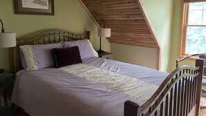 A bedroom with a wooden sloped ceiling, a bed with white bedding, two bedside lamps, and a framed picture on the wall.