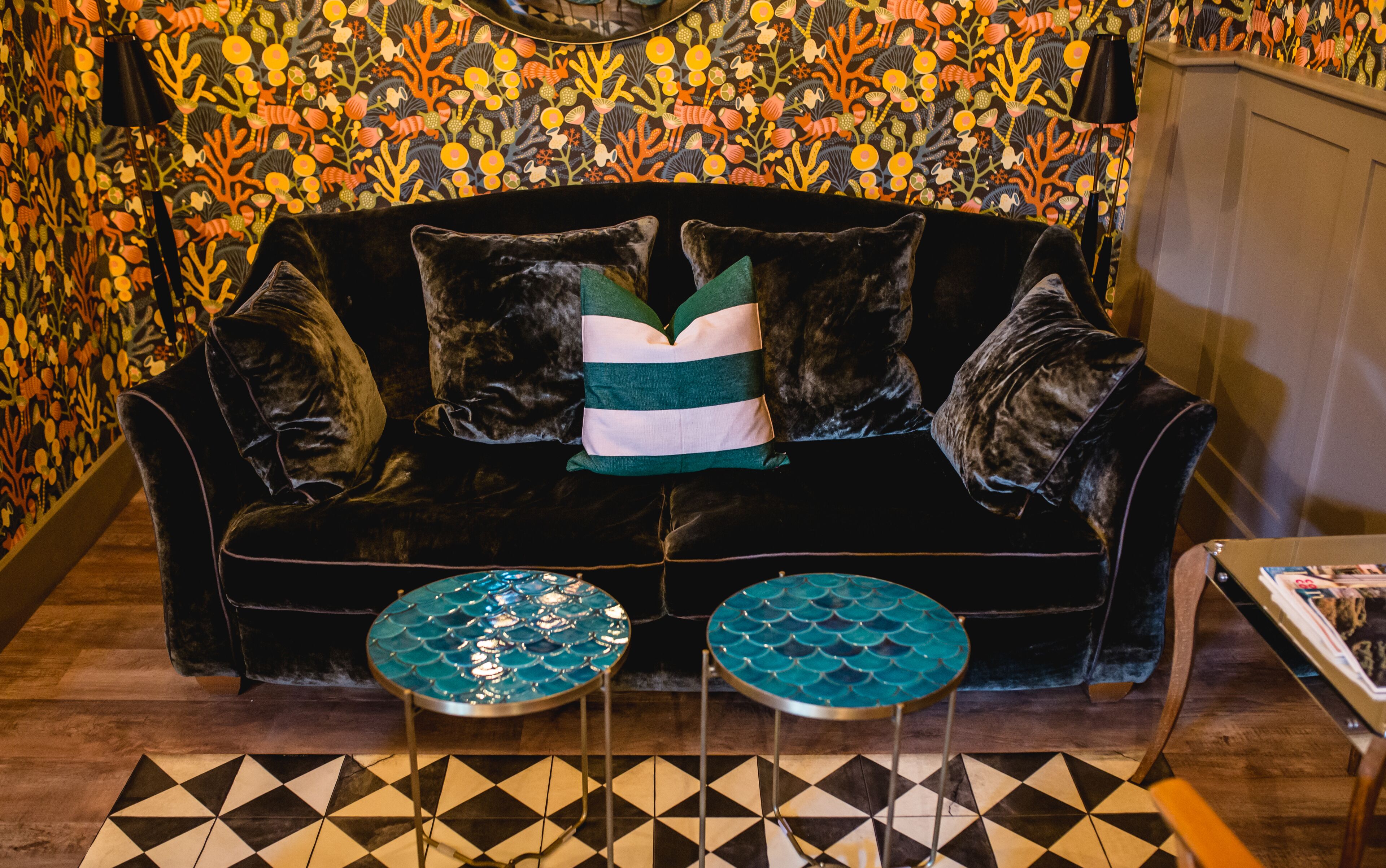 Un canapé en velours noir agrémenté d’un coussin à rayures vertes et blanches, deux tables de couleur turquoise ornées d’un motif en forme d’écailles de poisson, et un papier peint à motifs aux tons orange et jaune.