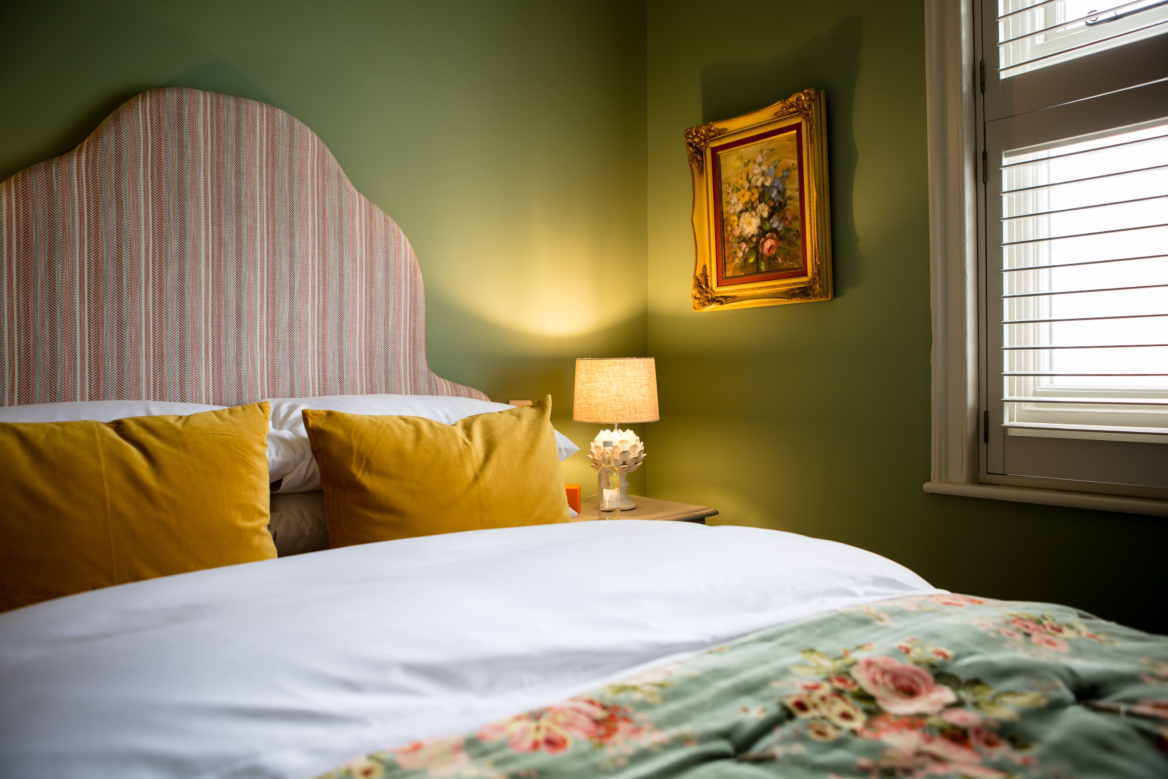 A bed with yellow pillows, a floral-patterned bedspread, a striped headboard, a bedside lamp, and a framed picture on the wall.