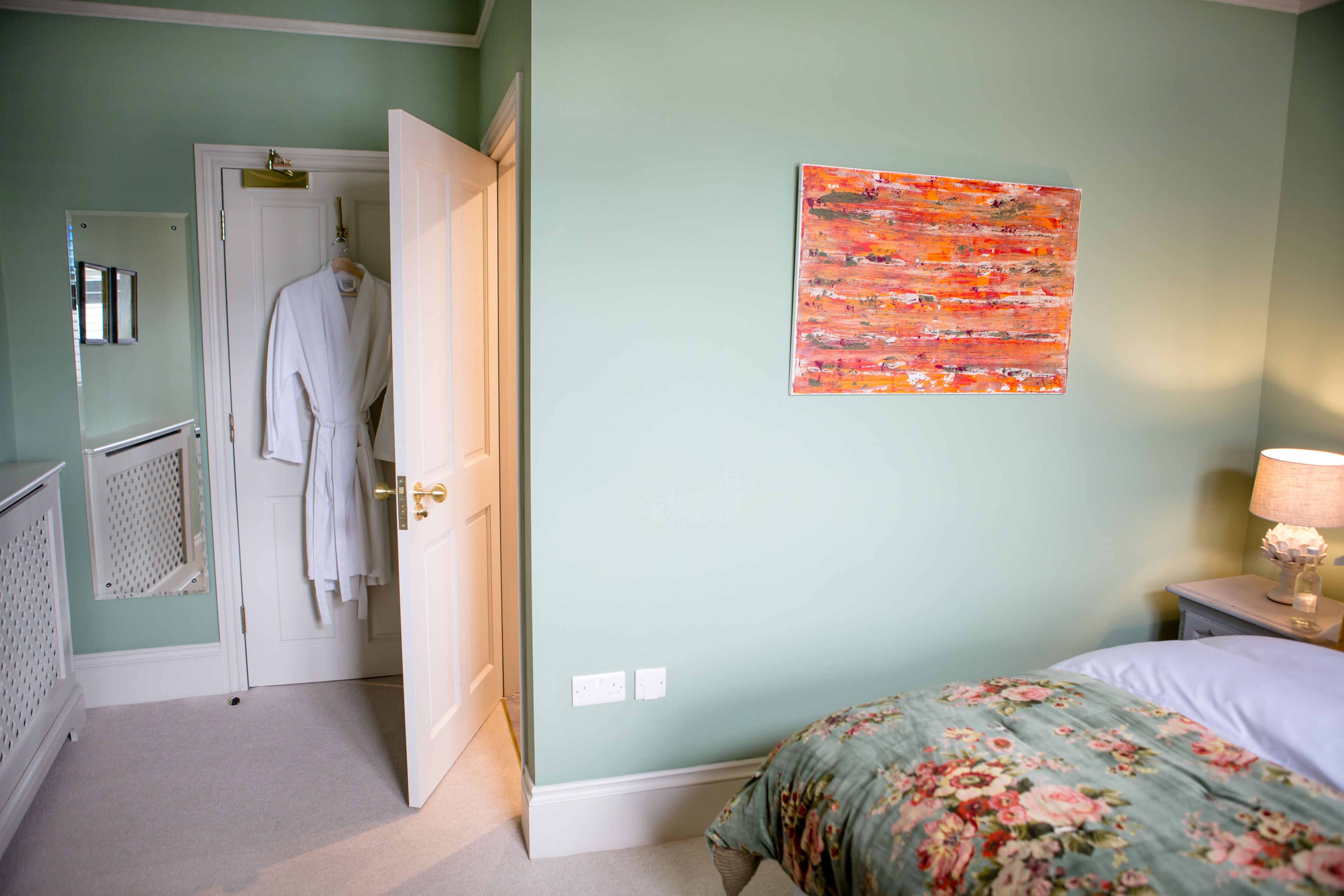 Un dormitorio con una cama, una mesita de noche, una obra de arte en la pared, una puerta y una bata colgada en un perchero.