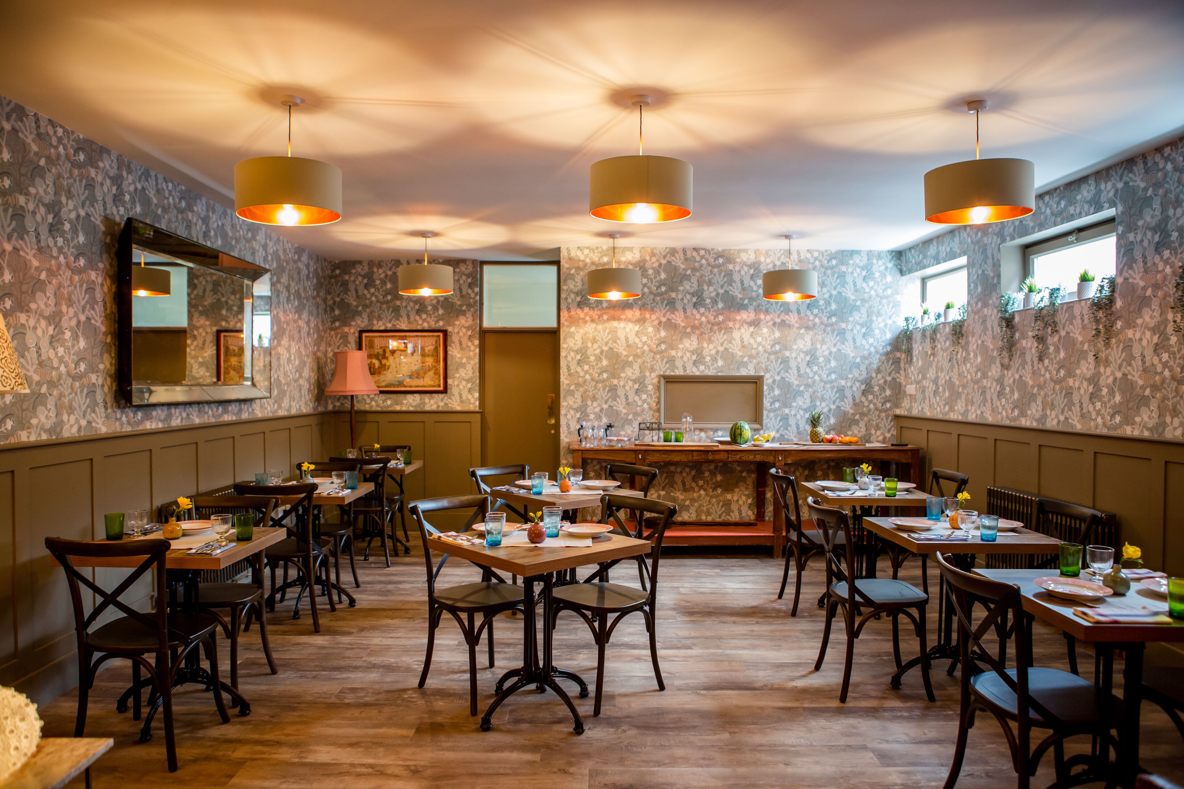 A restaurant with wooden tables and chairs, hanging pendant lights, and a bar area.