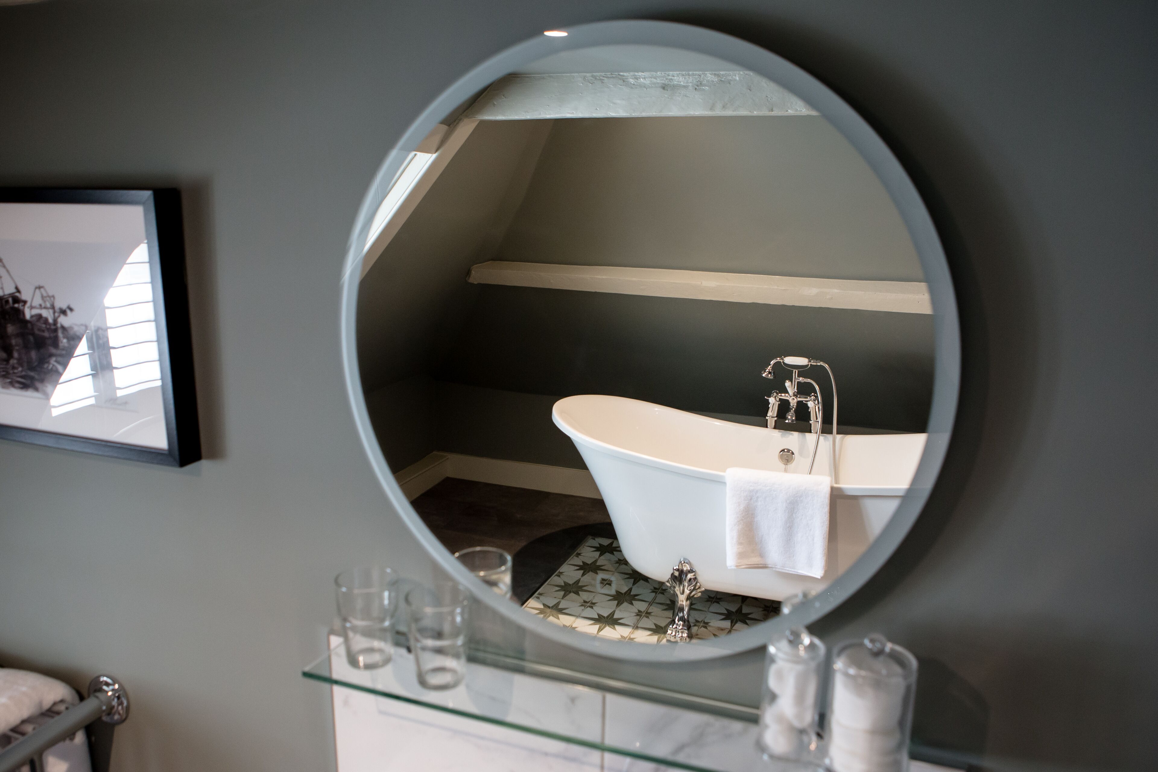 A bathroom with a freestanding bathtub, a round mirror, and a glass shelf with toiletries.