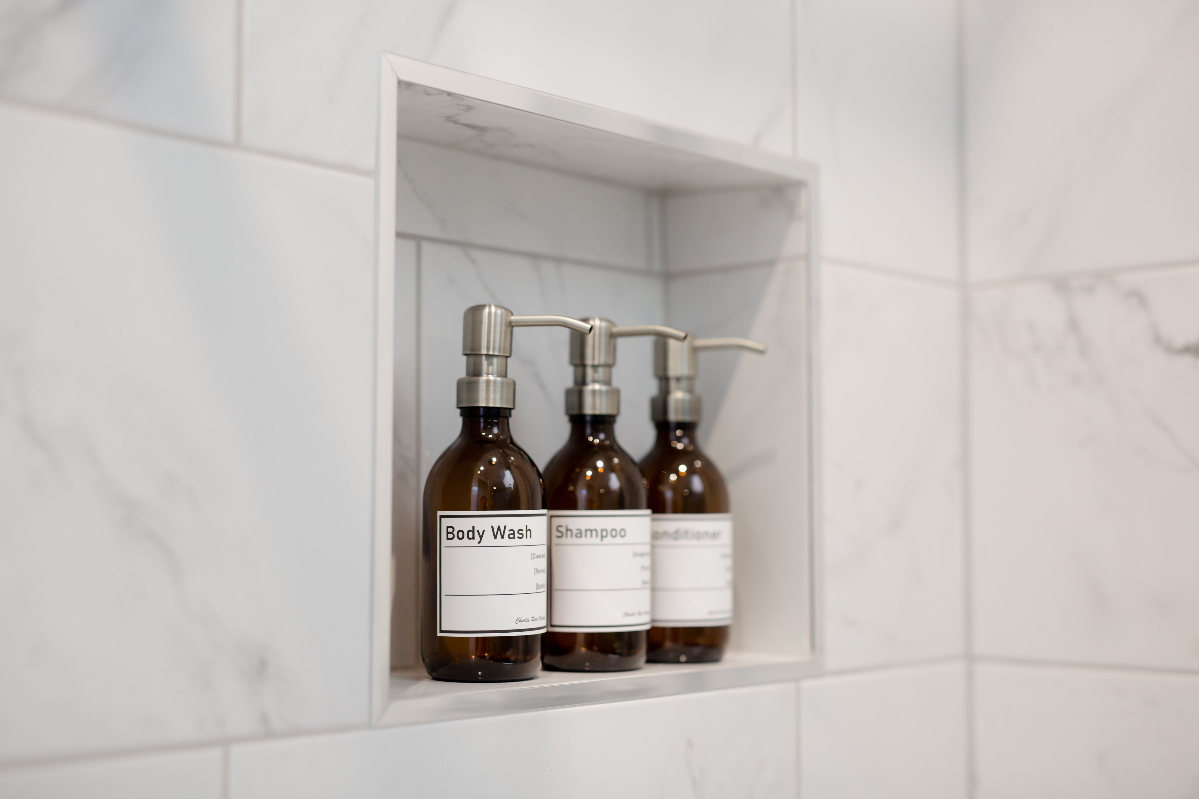 Three bottles of body wash and shampoo in a white shelf against a tiled wall.
