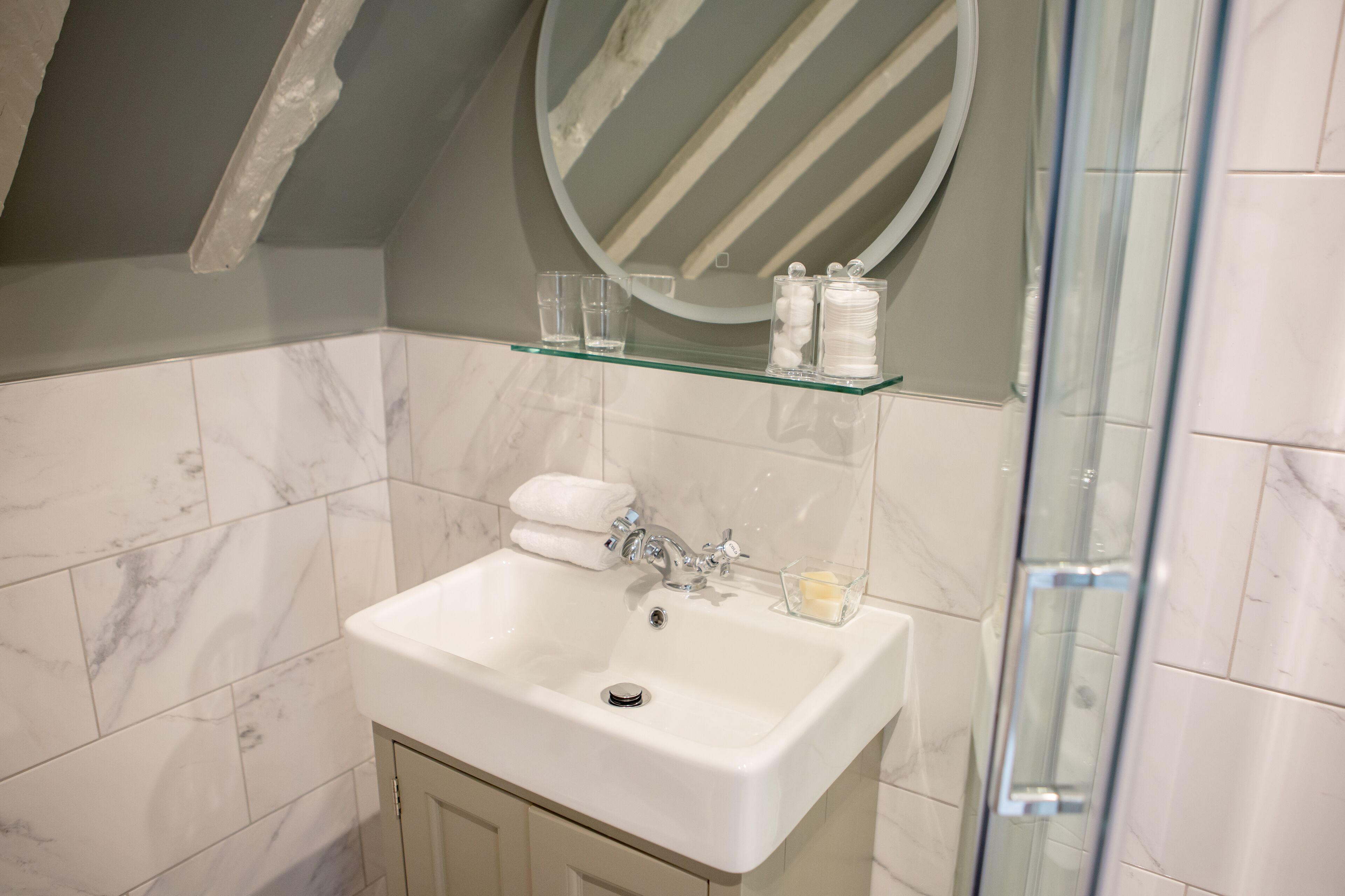 A bathroom with a white sink, marble tiles, a glass shelf with toiletries, and a round mirror.