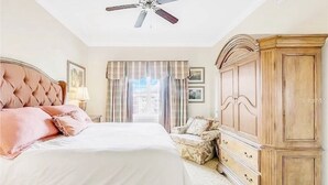 Un dormitorio con una cama grande, un ventilador de techo, una ventana con cortinas y un armario de madera.