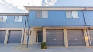 A two-story residential building with a blue exterior, two garage doors, and a small front yard with a bush. - Silvertrees - 2/1 McLure Cir (Jindabyne)
