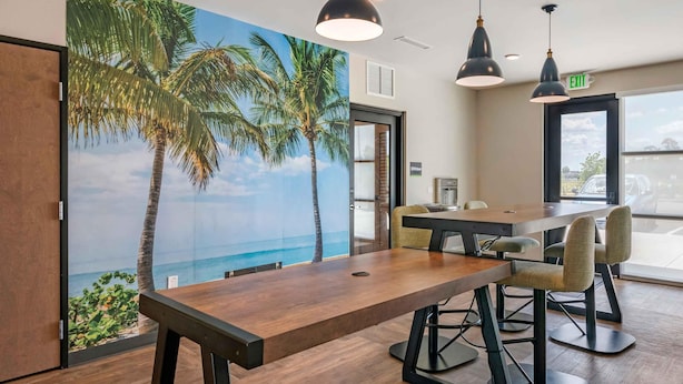 Een vergaderruimte met een houten tafel, stoelen, hanglampen en een muurschildering van palmbomen en een uitzicht op het strand.