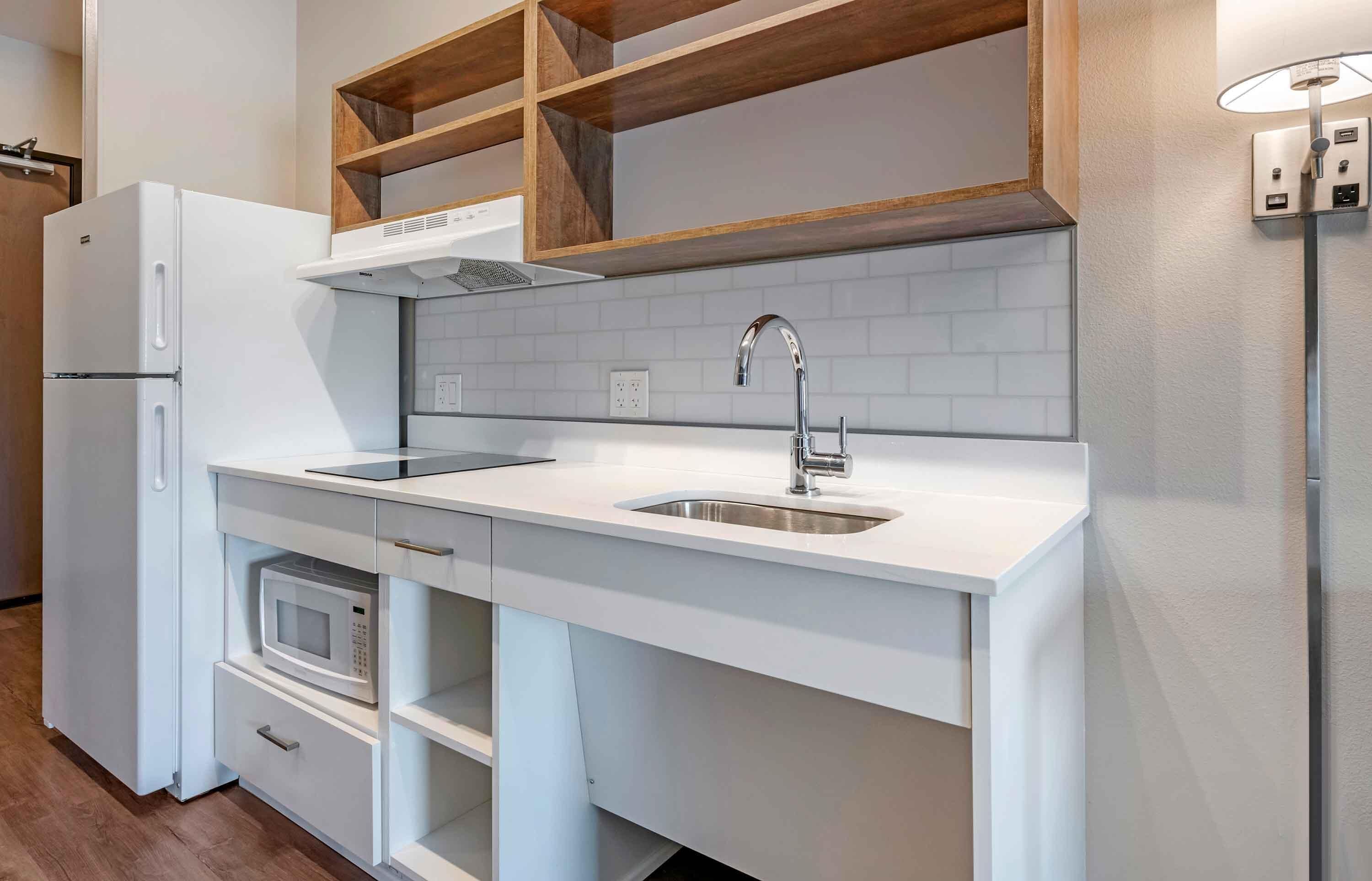 A modern kitchen with white cabinetry, a built-in microwave, a stainless steel sink, and wooden shelves above the countertop.
