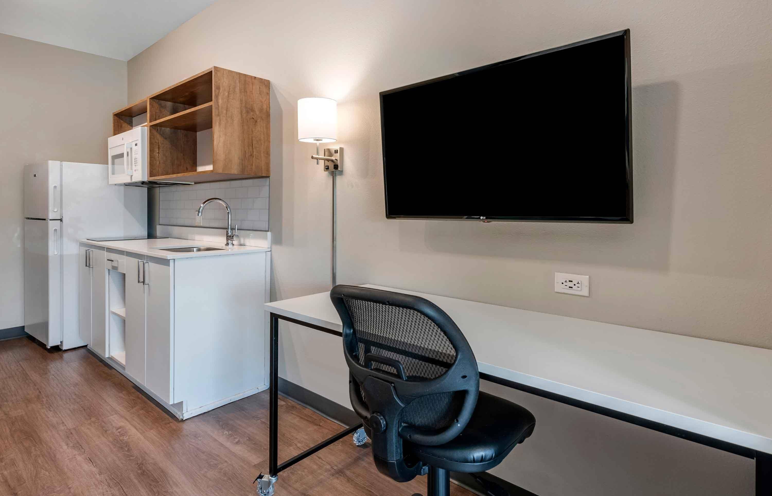 Een kitchenette met een magnetron, spoelbak en houten kastjes, een flatscreen televisie aan de muur en een bureau met stoel.
