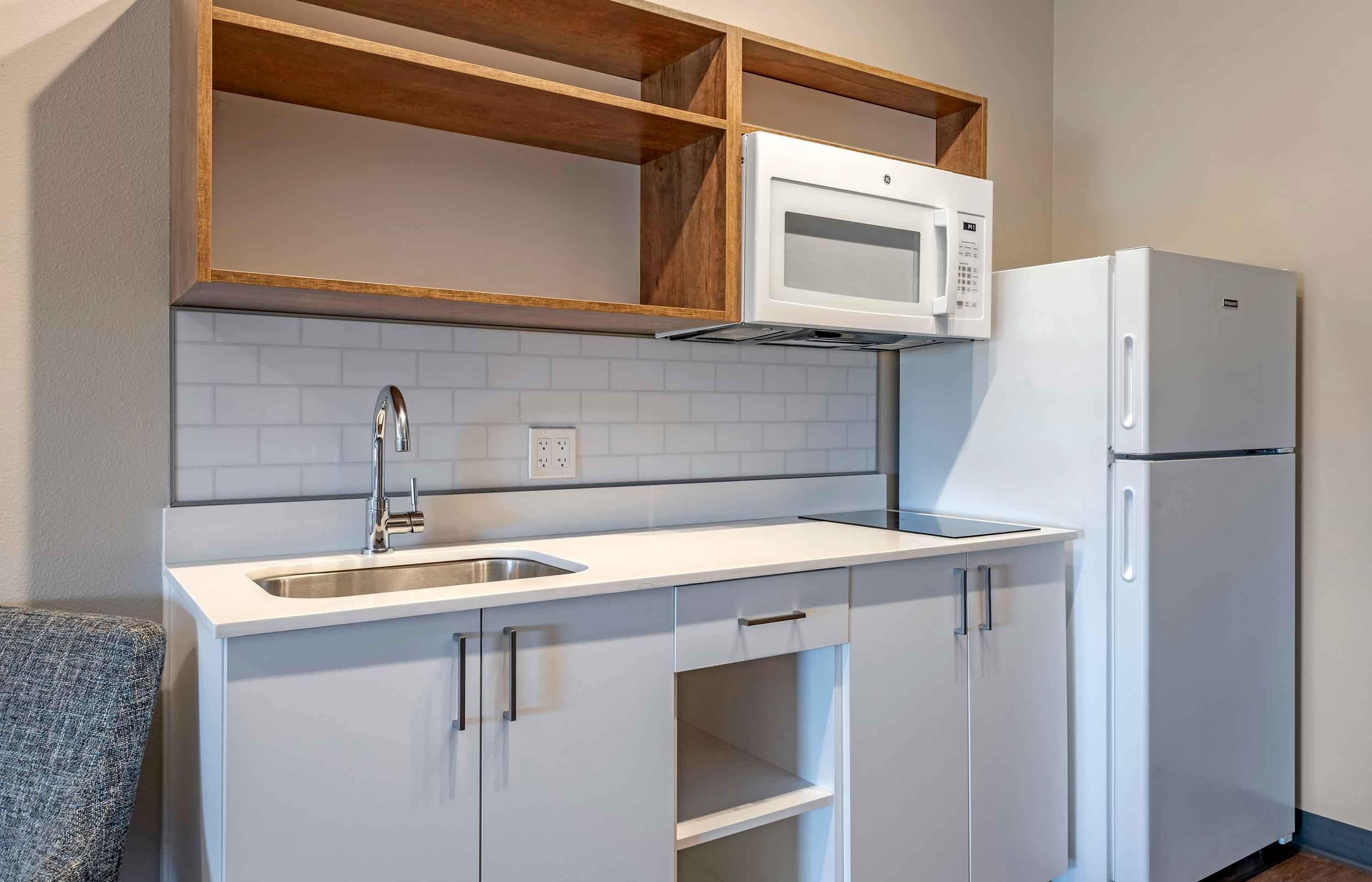Una cucina compatta con un piano di lavoro bianco, un forno a microonde integrato, un frigorifero ed alcune mensole in legno sopra il microonde.