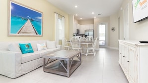 A spacious living room with a white sofa, a coffee table, and a dining area with white chairs. A kitchen is visible in the background, and a large framed picture of a beach scene is on the wall. - Relax & Reboot - Fantastic Margaritaville Cottage w/ Private Hot Tub (Kissimmee)
