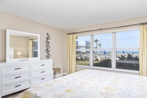 A bedroom with a large bed, a dresser, a mirror, and a window with a view of the beach and buildings. - Sandpiper 103 - A Few Steps to Beach (St. Augustine)