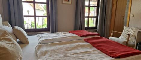 Ein Hotelzimmer mit zwei Betten, einem Sessel, einem Schreibtisch und großen Fenstern mit Vorhängen.