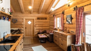 A log cabin interior with a wooden ceiling, a dining area with a table and benches, a kitchen with a sink and wooden cabinets, and a window with checkered curtains. - Ellerbeck Gesundheits- und Wellnesshof (Langdorf)