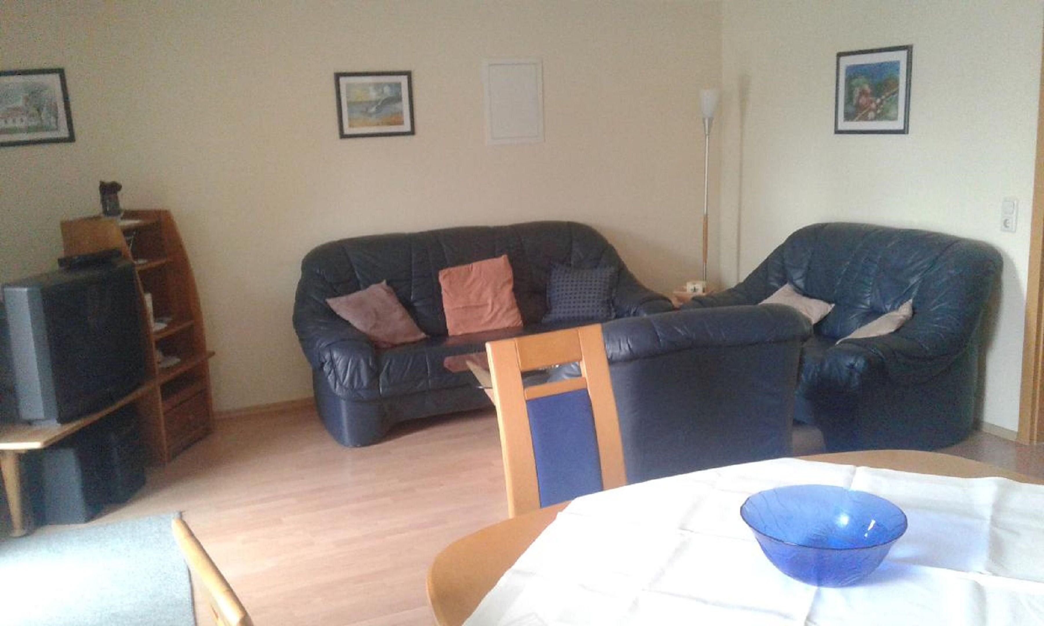 A living room with a black leather sofa, a wooden chair, a dining table with a blue bowl, and a television on a wooden stand.