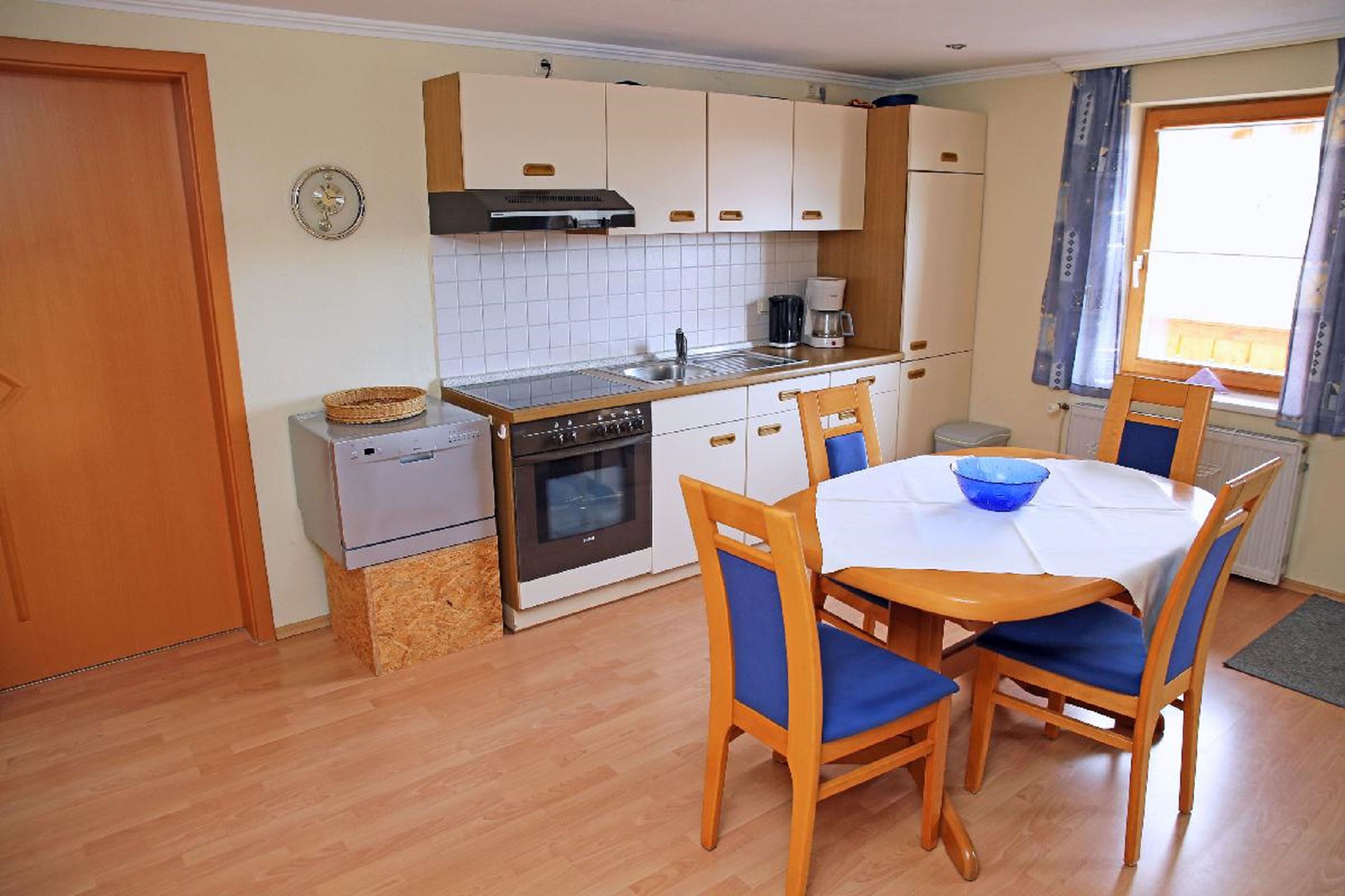 A compact kitchen with a stove, oven, and sink, a dining table with chairs, and a window with curtains.
