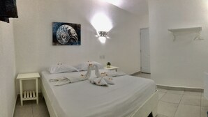 Quarto de hotel com cama bem arrumada, uma obra de arte na parede, uma mesa de cabeceira com abajur e uma mesinha lateral.