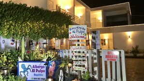 Entrada de hotel com uma placa de boas-vindas, uma placa indicando 'Aibergu El Pulpo Bayahibe' e um aviso de que o hotel está aberto 24 horas.