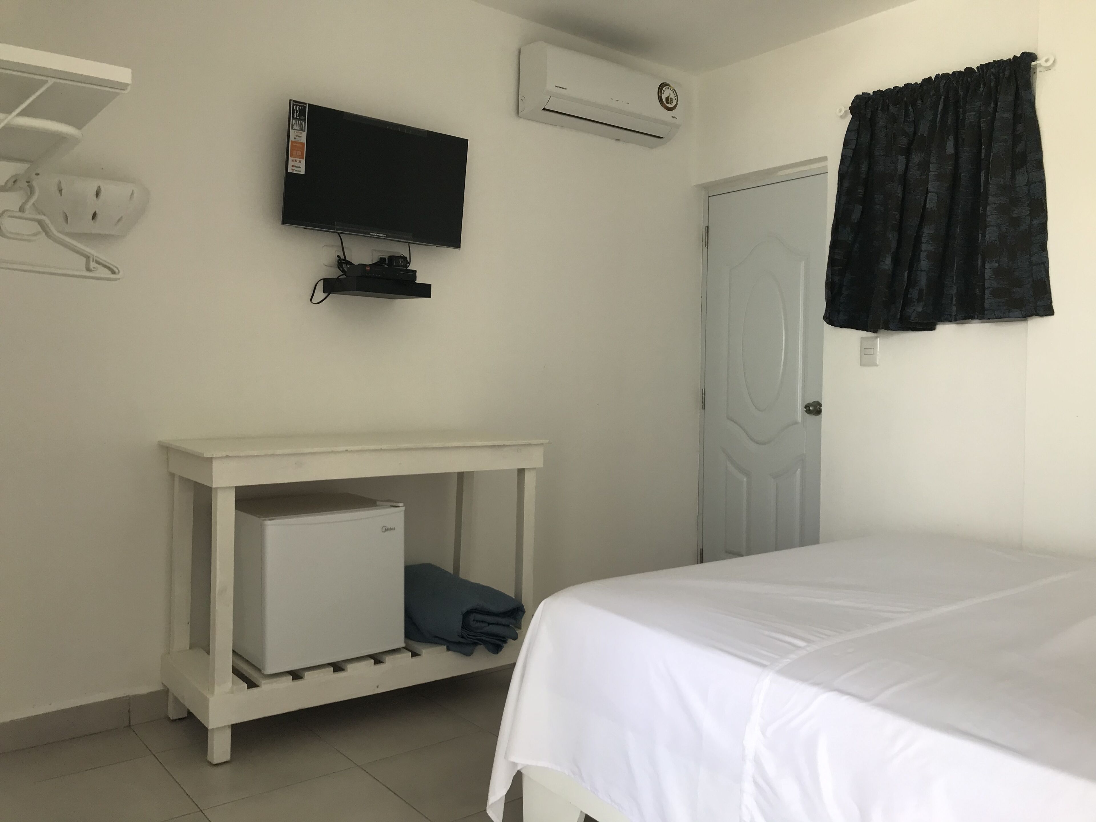 A bedroom with a bed, a TV mounted on the wall, a small white cabinet, and a door with a curtain.