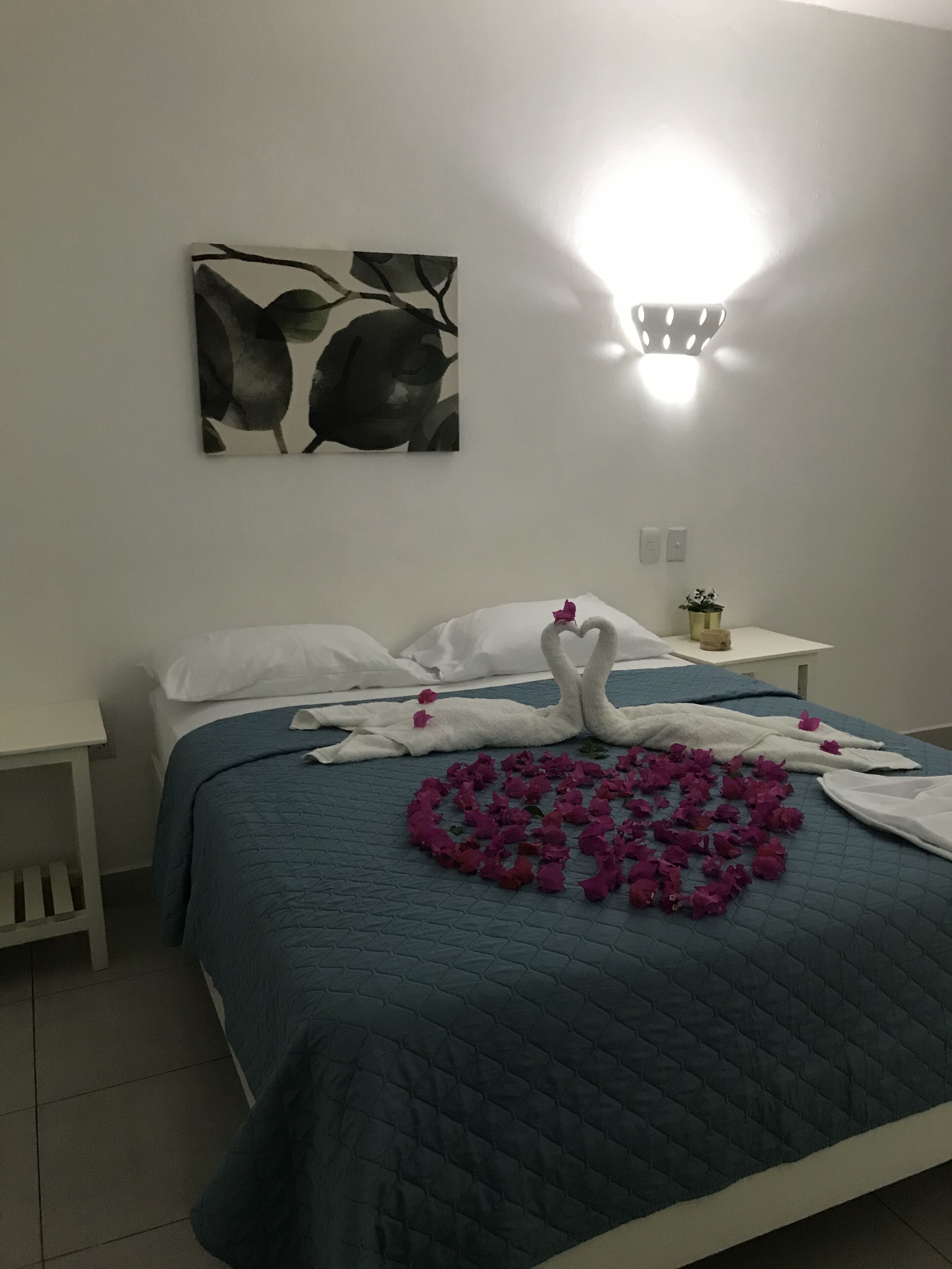 Un letto rifatto con una coperta blu, cuscini bianchi e un asciugamano disposto a forma di cuore con fiori rosa su un pavimento piastrellato.