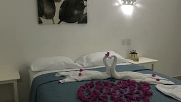 A neatly made bed with a blue quilt, white pillows, and a towel arranged in a heart shape with pink flowers on a tiled floor.