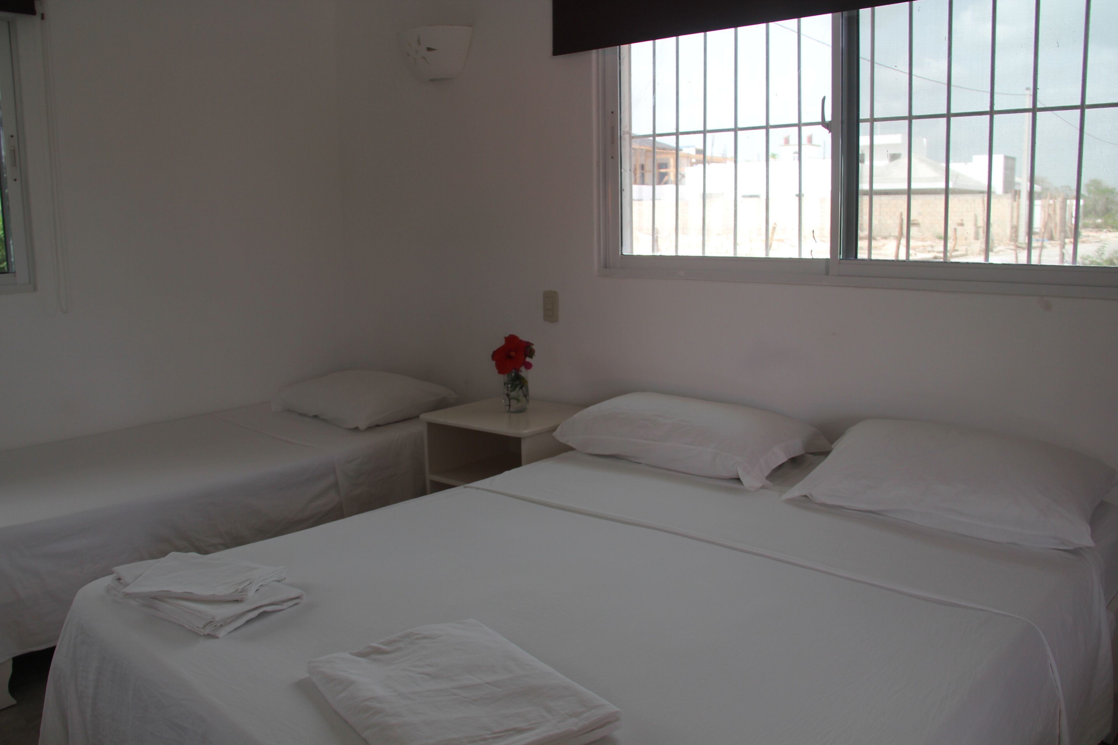 A hotel room with two single beds, a small table with a vase of red flowers, and a window with a view of buildings outside.