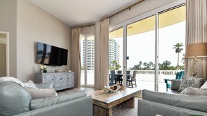 Una sala de estar moderna con una televisión de pantalla plana, sofás grises, una mesa de centro de madera y vistas a un rascacielos y palmeras a través de amplias ventanas.