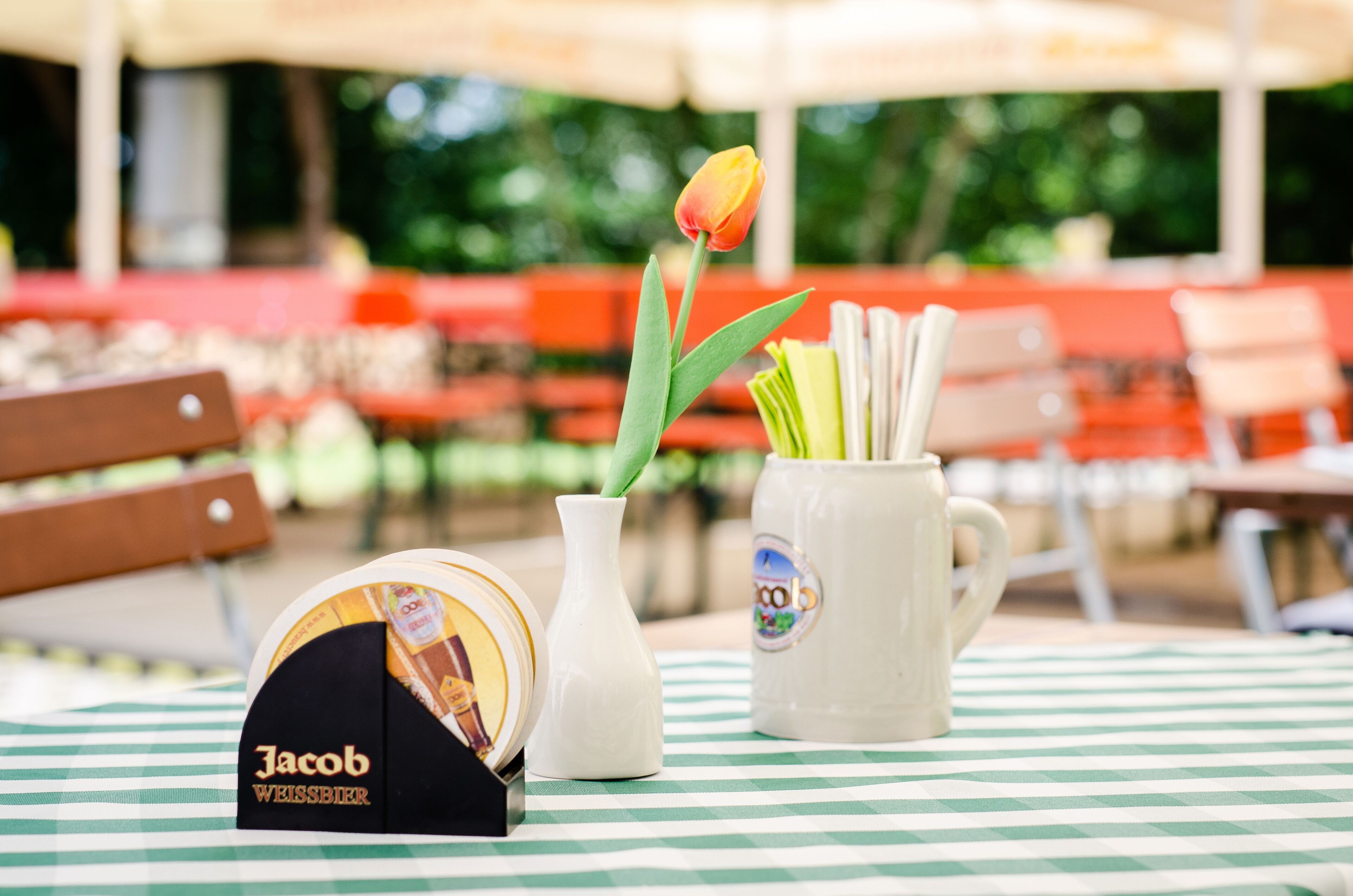 A table with a checkered tablecloth, a white mug with a label, a white vase with a tulip, and a box labeled "Jacob Weissbier".