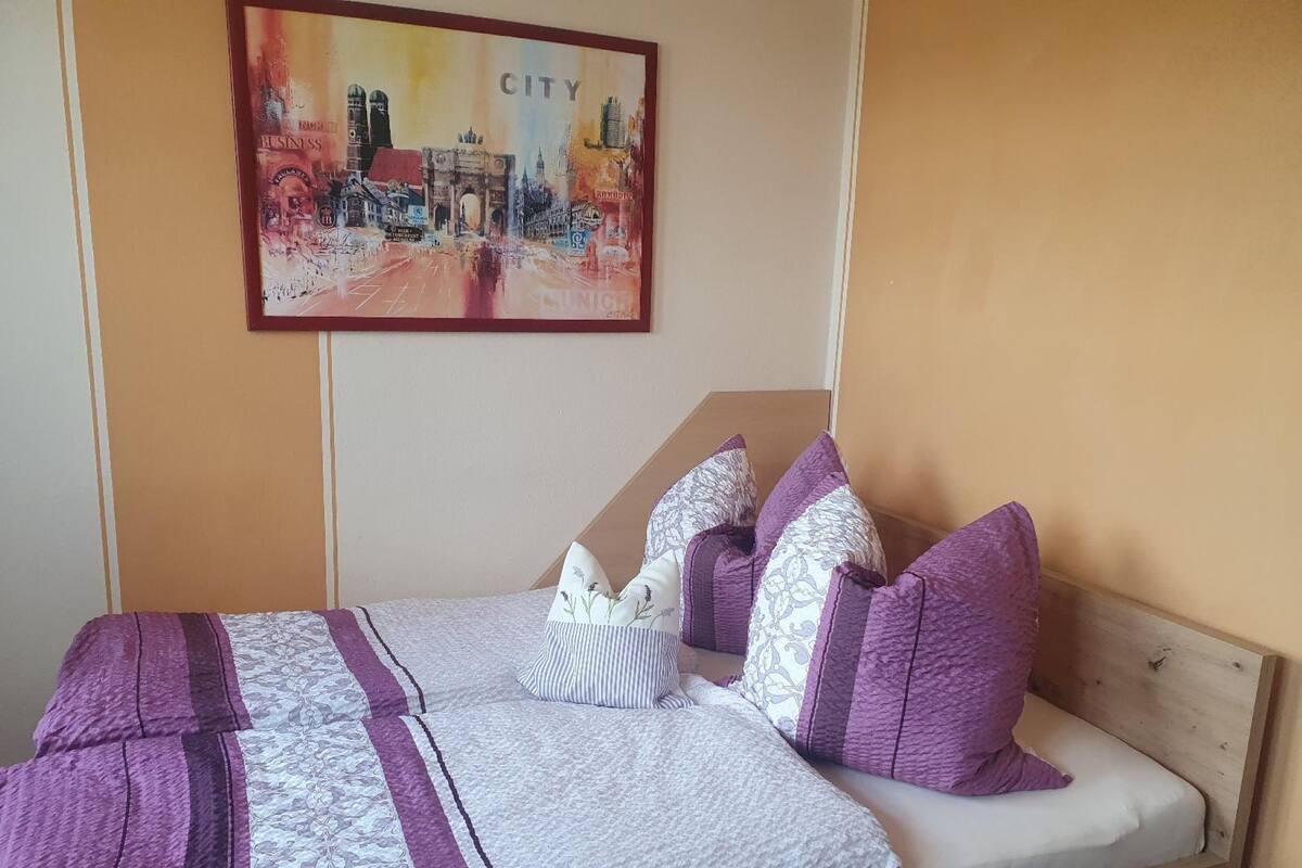 A bed with a patterned bedspread, multiple pillows, and a cityscape painting on the wall.