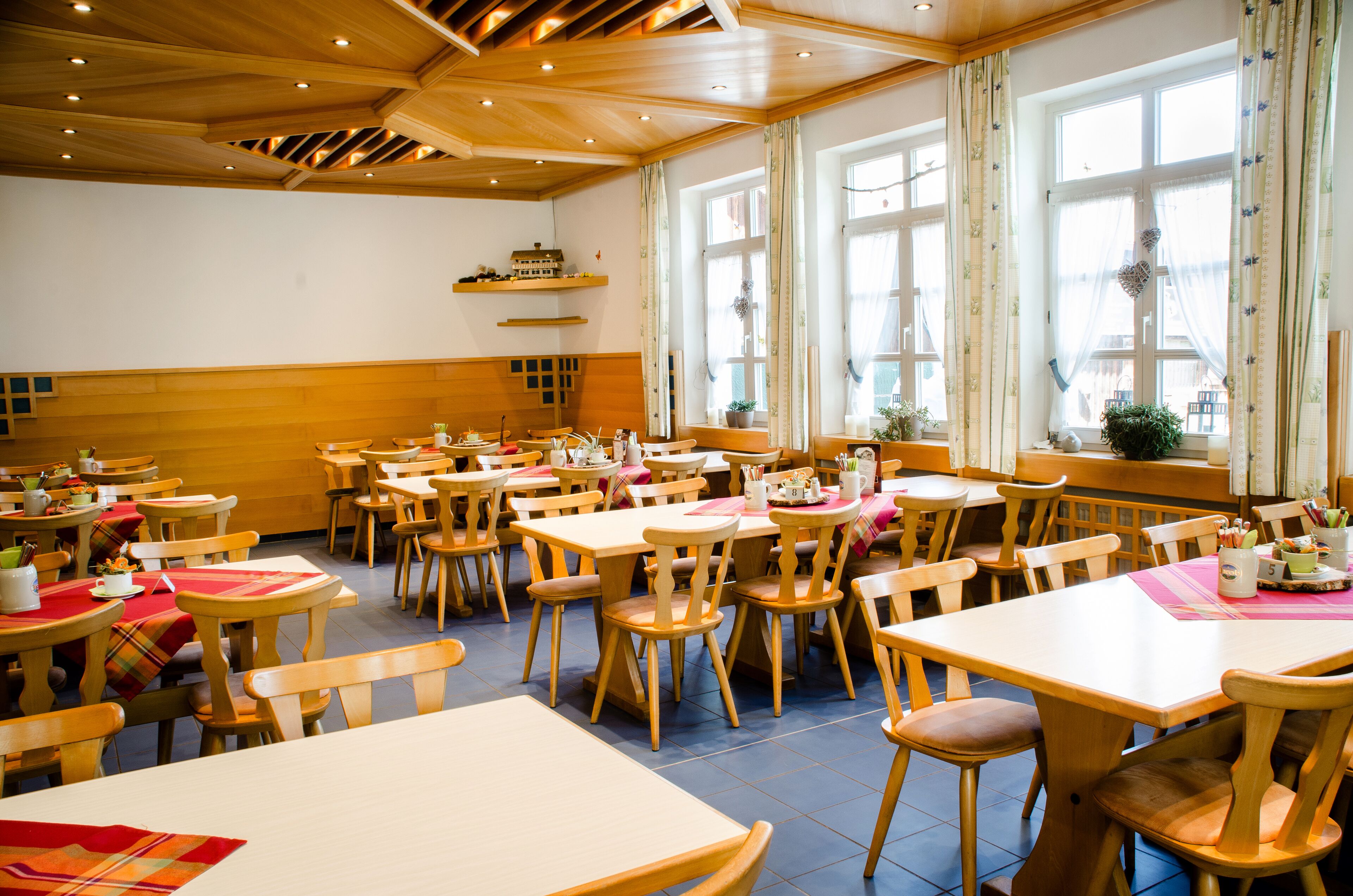 Un restaurante con mesas y sillas de madera, amplias ventanas con cortinas y un techo de madera con iluminación empotrada.