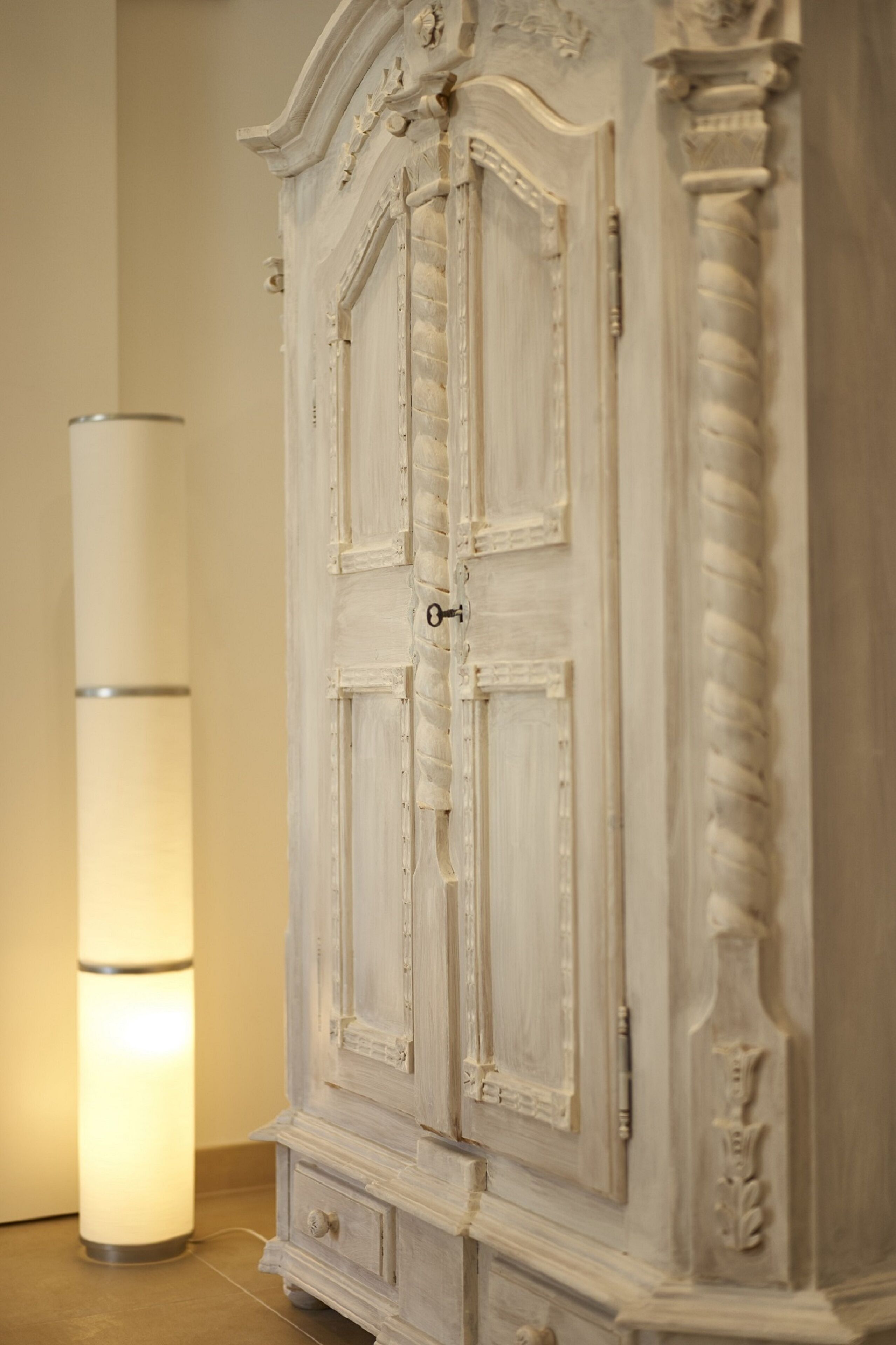 A white, ornate cabinet with a keyhole and a drawer, next to a tall, cylindrical, illuminated lamp.