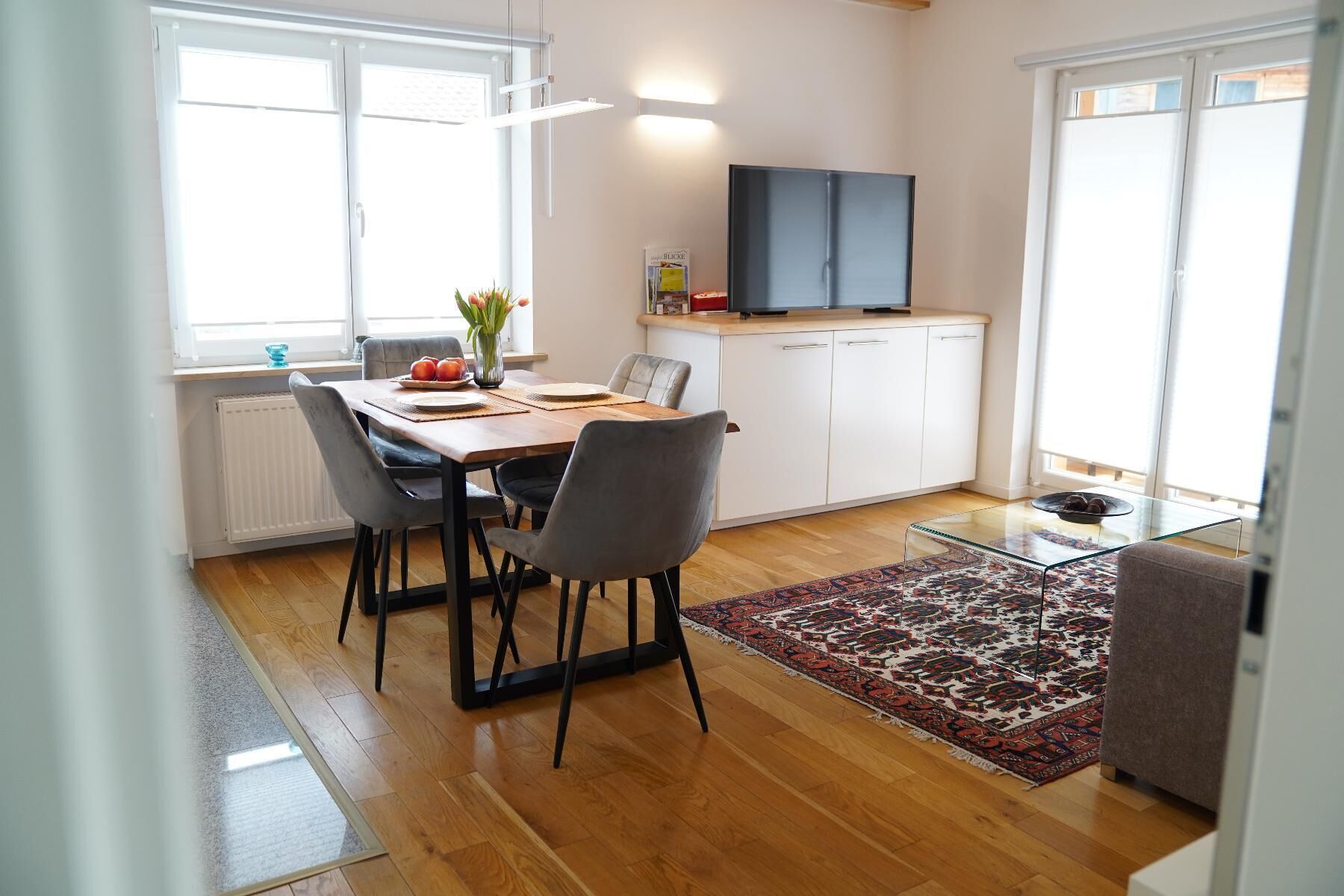 A modern kitchen with a dining area, wooden flooring, and a large window providing natural light.