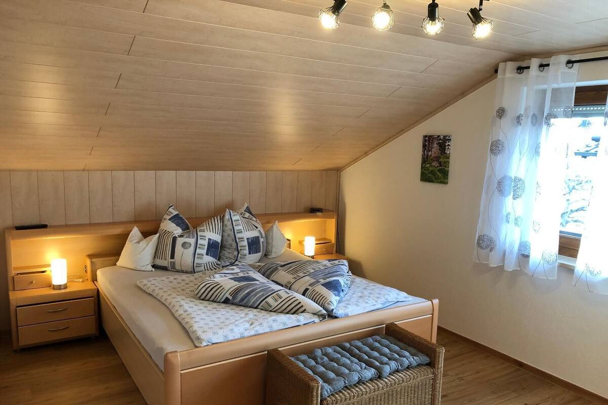 Un dormitorio con techo inclinado, cabecera de madera, cama con ropa de cama estampada, lámparas de noche, una cesta de mimbre y una ventana con cortinas traslúcidas.