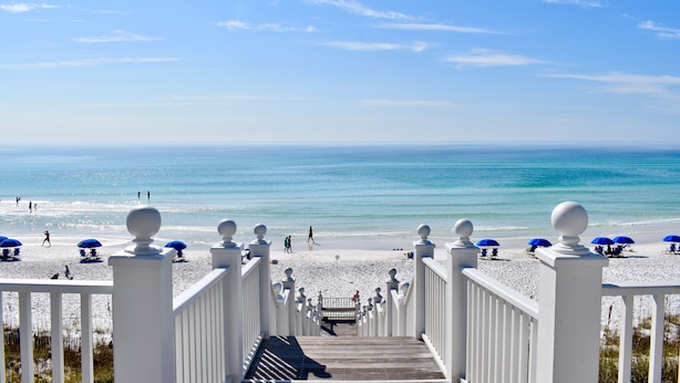 Een strand met een houten looppad dat naar het strand leidt, blauwe parasols en witte leuningen.