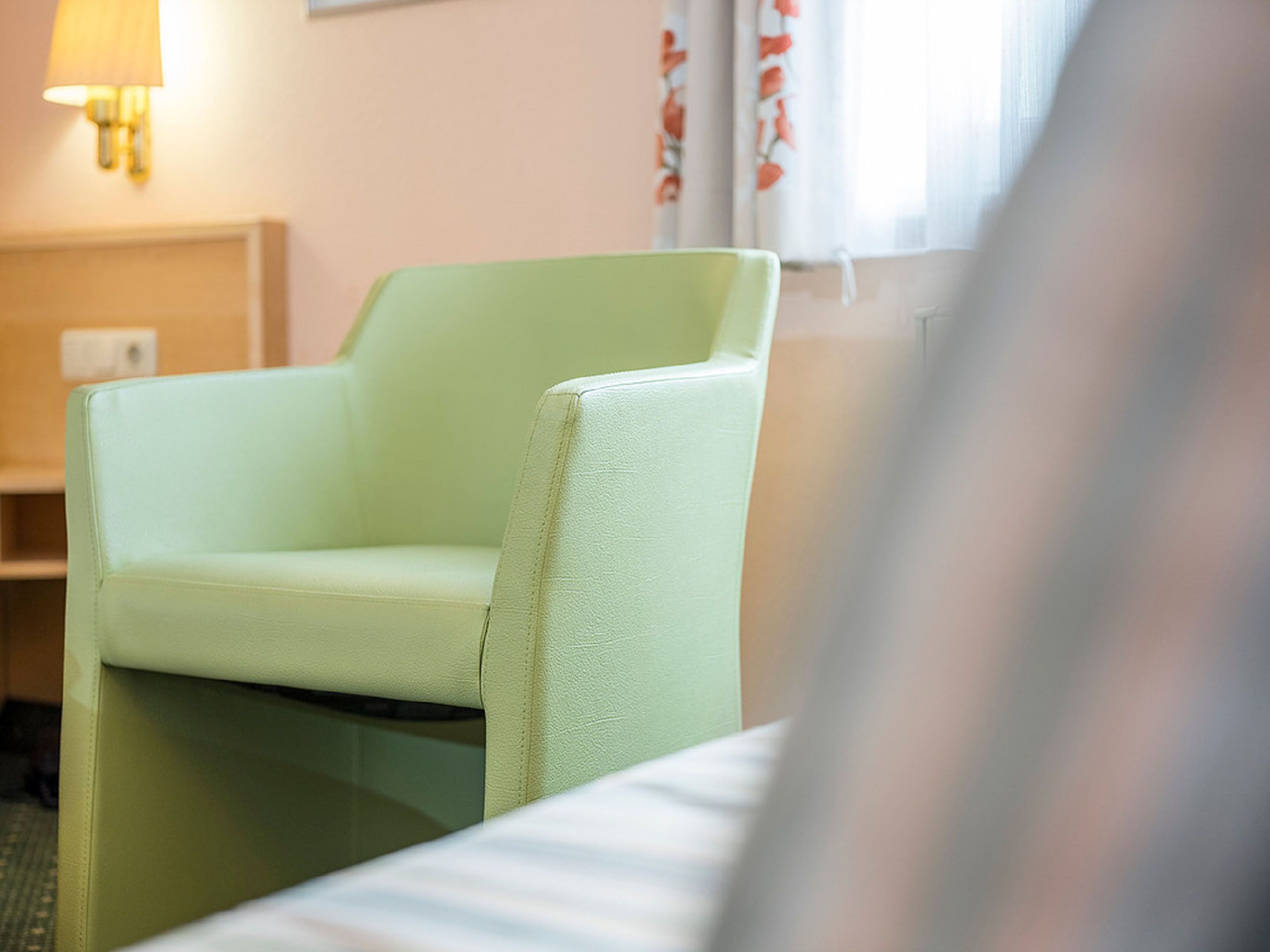 A light green armchair in a hotel room with a bedside table and a lamp.