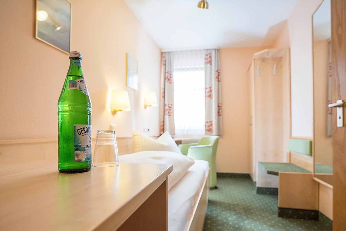 Een hotelkamer met een bed, een nachtkastje met fles en glas, een stoel, een bureau, een spiegel en een douchehoek.
