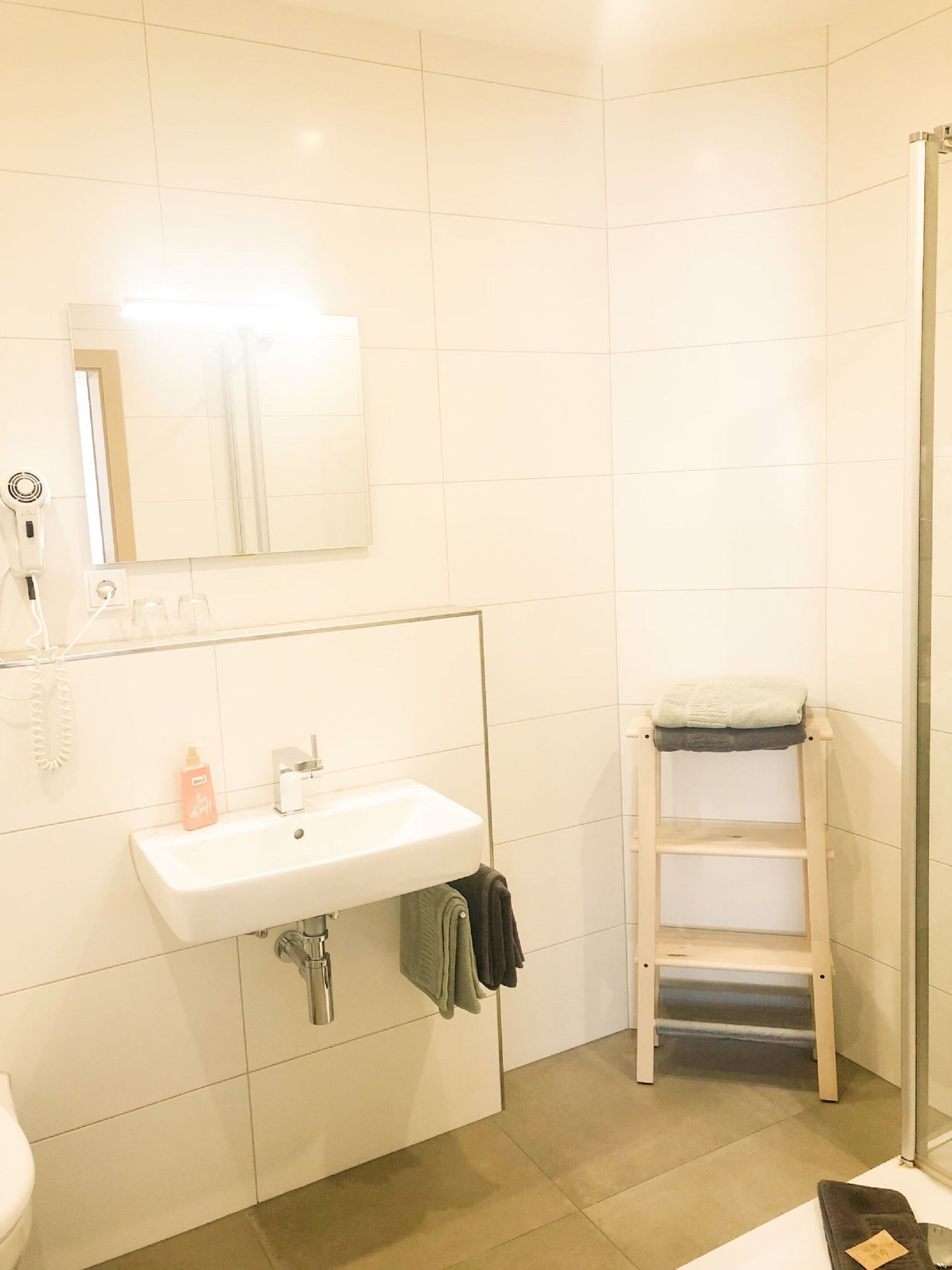 Baño con lavamanos blanco, espejo y ducha. Se aprecia un perchero con una toalla y un pequeño estante con una botella.