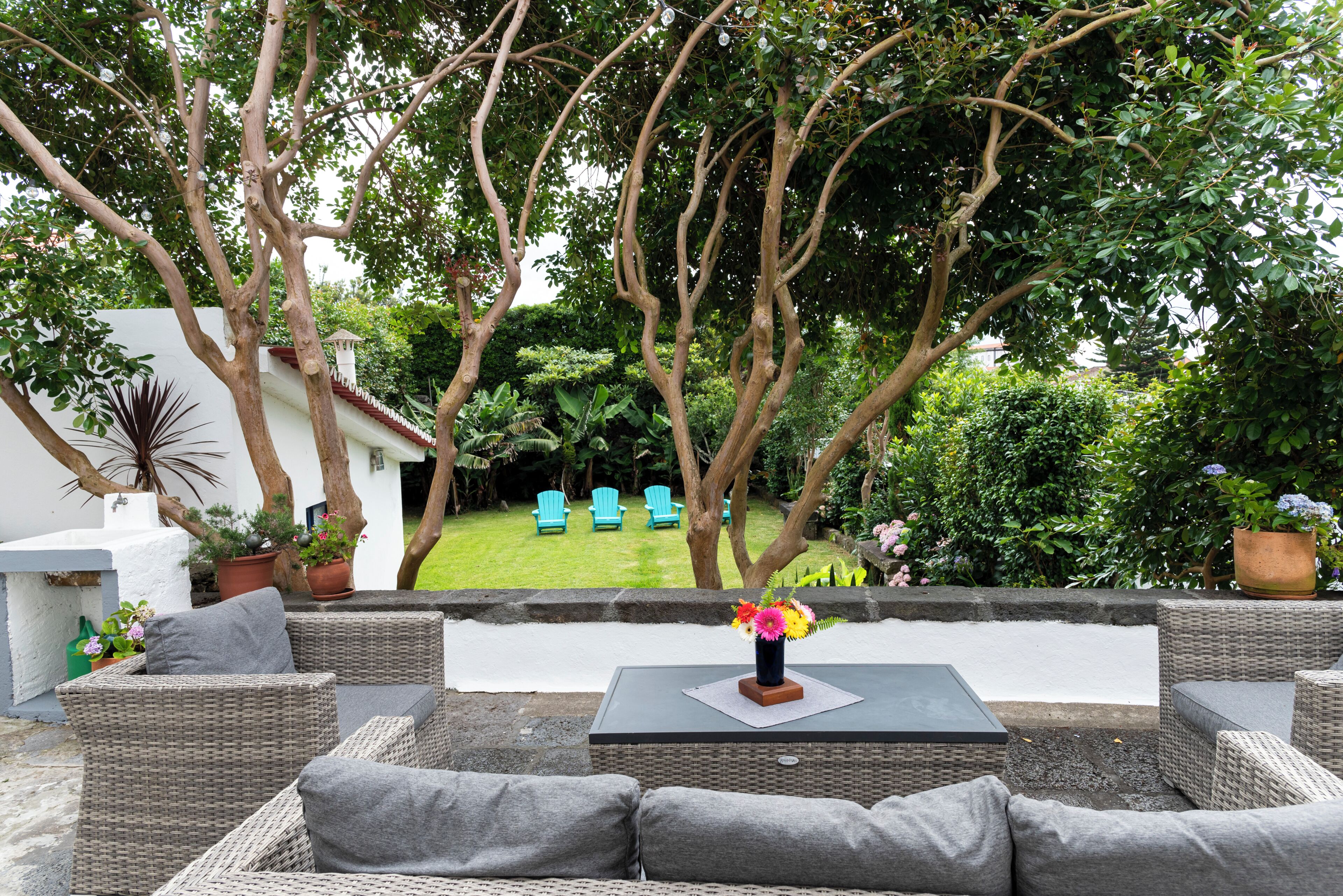 Eine Terrassenfläche mit Korbmöbeln, einem Tisch mit Blumenvase und Blick auf einen Garten mit Bäumen und Stühlen.