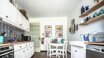 Une cuisine avec des meubles blancs, un plan de travail en bois et un coin repas avec des chaises blanches. Des tableaux encadrés ornent le mur, et un extincteur est placé près de la porte.